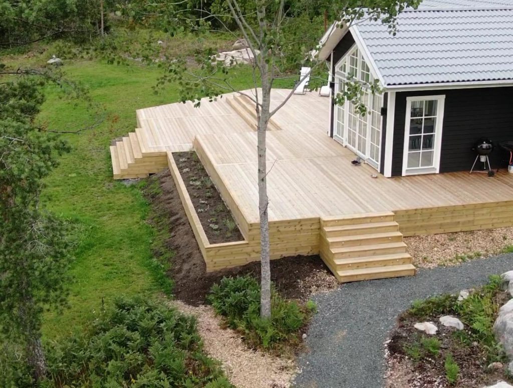 Bilden visar en stor träaltan vid ett svart hus med vita detaljer. Altanen har flera nivåer, en blomlåda längs kanten och en trappa som leder ner till trädgården.